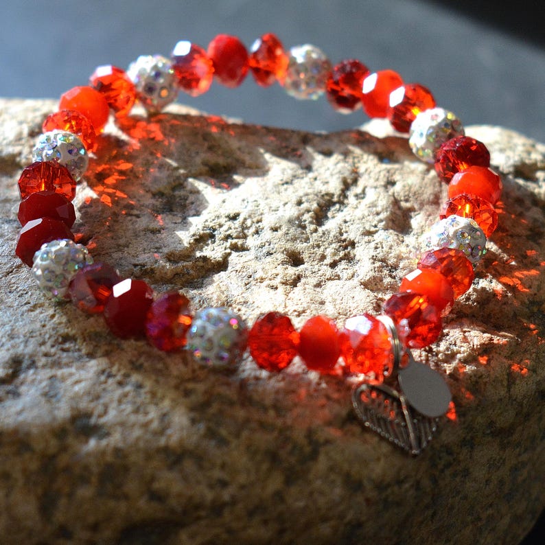 Puede incluir: Una pulsera roja con cuentas facetadas y cuentas plateadas con incrustaciones de pedrer&iacute;a. La pulsera tiene un dije de hoja plateada y se muestra sobre una superficie de piedra texturizada.