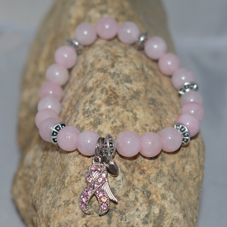 Op de afbeelding: Roze rozenkwarts kralen armband met een zilveren bedel met een roze lint met strass. De armband ligt op een stenen achtergrond.