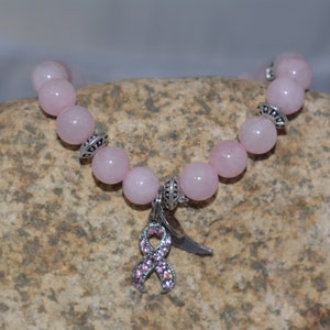 Op de afbeelding: Een armband van roze edelstenen met een zilveren bedel in de vorm van een roze lint met strass. De armband ligt op een bruine steen.