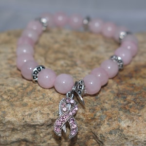 Op de afbeelding: Een roze rozenkwarts kralen armband met een zilveren bedel met een roze lint met strass. De armband ligt op een bruine rotsachtige ondergrond.