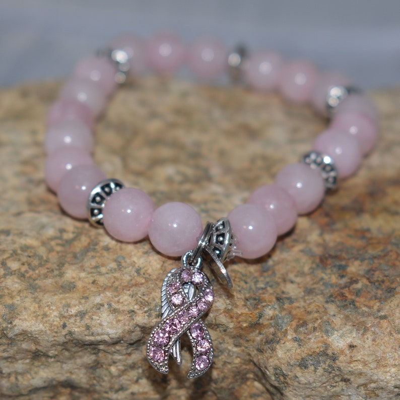 Op de afbeelding: Een roze rozenkwarts kralen armband met een zilveren bedel met een roze lint met strass. De armband ligt op een bruine rotsachtige ondergrond.