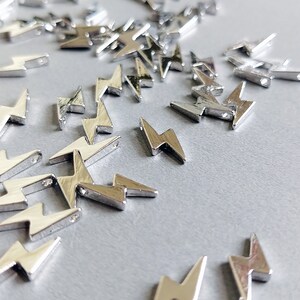 Op de afbeelding: Een verzameling kleine, zilverkleurige bliksembehang. Elke bedel heeft een driedimensionaal ontwerp met een gat aan de bovenkant om te rijgen. De bedels zijn verspreid over een lichtgrijze ondergrond.