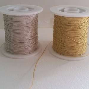 May include: Two spools of metallic thread, one silver and one gold. The thread is wound around white plastic spools. The silver thread has a subtle shimmer, while the gold thread is bright and shiny. The spools are on a white surface. The thread is suitable for crafts.