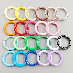 Puede incluir: Una colección de cierres redondos de metal de colores en varios tonos, incluyendo amarillo, naranja, rojo, rosa, verde, azul, blanco, negro y tonos metálicos. Estos cierres están diseñados para manualidades o joyería.