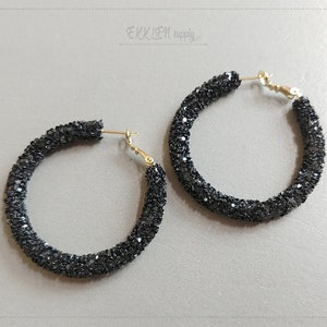 May include: A pair of black glitter hoop earrings with gold-colored clasps. The earrings are round and covered in small, sparkling black glitter. The background is a light gray surface.