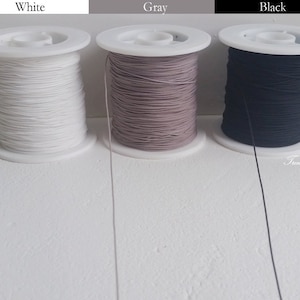 May include: Three spools of elastic cord in white, gray, and black. Each spool is wound on a white plastic base. The text "White", "Gray", and "Black" are above each spool. The words "Trend Tassel" are in the bottom right corner.