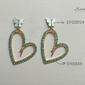 May include: A pair of heart-shaped earrings with a gold-colored frame, adorned with small, light green beads and clear crystals. Each earring features a light blue butterfly at the top. The earrings are labeled "EPG0054" and "EH0049".