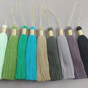 May include: A selection of nine decorative tassels in a range of colours. Each tassel has a gold-coloured metallic cap and a loop for hanging. The colours include green, blue, grey, and black.