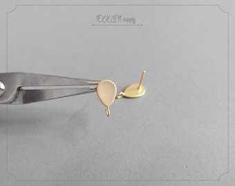 May include: Close-up of gold-colored earring components. One earring component is held by pliers, showing a teardrop-shaped charm with a small hook. The other earring component is a post with a round base. The background is a light gray.