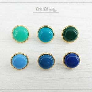May include: Six round cabochon buttons in shades of teal, turquoise, green, and blue. Each button has a gold-colored, textured rim. The buttons are arranged in two rows of three on a white surface. The text "EKKLEN supply" is in the upper right corner.