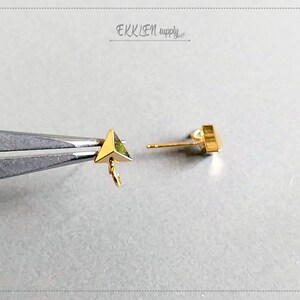 May include: Close-up of gold-colored triangle stud earrings. One earring is held by tweezers, showcasing a small, geometric design with a loop for attaching charms. The other earring is a post-style stud with a square shape.