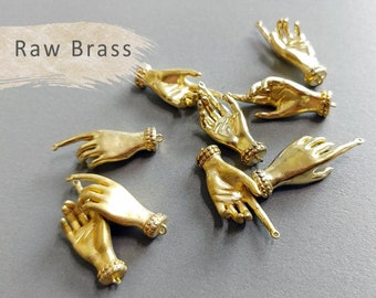 May include: Several small, gold-colored brass hand charms with various finger positions. Each charm has a detailed wrist cuff and a small loop for attaching to jewelry. The text "Raw Brass" is in the upper left corner.