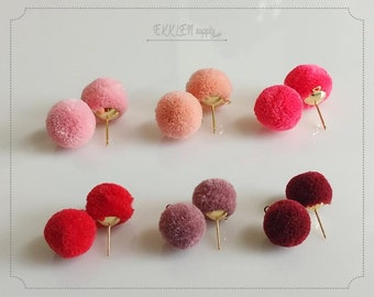 May include: A collection of pom-pom stud earrings in various shades of pink, peach, red, and burgundy. Each pair features a fluffy, round pom-pom attached to a gold-colored post. The earrings are displayed on a white surface.