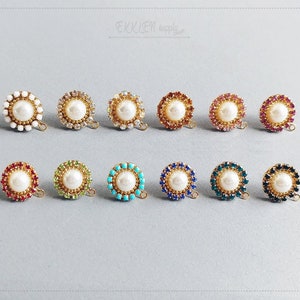 Puede incluir: Una colección de doce componentes de joyería redondos. Cada componente presenta una perla blanca central rodeada por un anillo de pedrería de colores. La pedrería es de varios colores, incluyendo rojo, verde, azul y dorado. Los componentes tienen un lazo dorado.