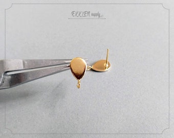 May include: Close-up of two gold-colored teardrop-shaped earring components. One is held by silver pliers, the other is on a light gray surface. Each earring component has a post and a small loop at the bottom.
