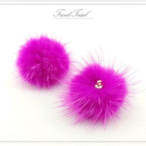 May include: Two bright pink fluffy pom poms with a gold button in the center of each.