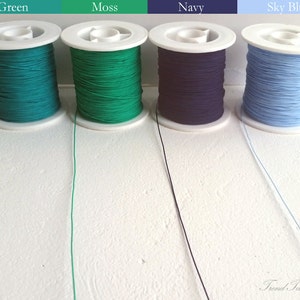 May include: Four spools of thread in shades of green, moss, navy, and sky blue. Each spool is white with the thread wound around it. The colors are labeled with text above each spool. The text "Trend Tassel" is in the bottom right corner.