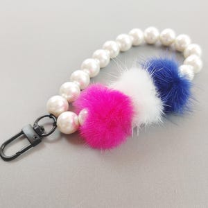 Op de afbeelding: Een polsbandje sleutelhanger met een streng witte imitatieparels, een zwarte sluiting en drie pluizige pompons in roze, wit en blauw. De sleutelhanger is ontworpen om aan sleutels of tassen te bevestigen.