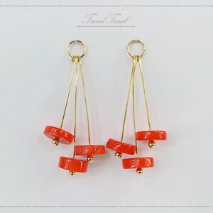May include: Dangle earrings with a gold-tone metal design. Each earring has a gold hoop, with three gold wires holding three bright orange, circular beads. The text "Trond Tassel" is in the upper center. The earrings are a unique accessory.
