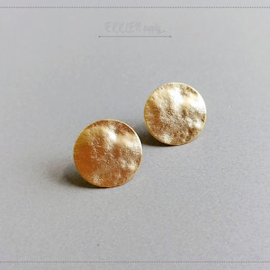 May include: Two gold-colored, textured, round stud earrings. The earrings have a hammered metal appearance and are displayed on a light gray surface. The earrings are a simple, elegant jewelry item.