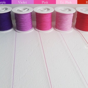 Puede incluir: Cinco carretes de hilo en fila, cada uno con un color diferente: morado, violeta, rosa, rosa claro y rojo. Los carretes son blancos con el hilo enrollado. Los colores están etiquetados encima de cada carrete.