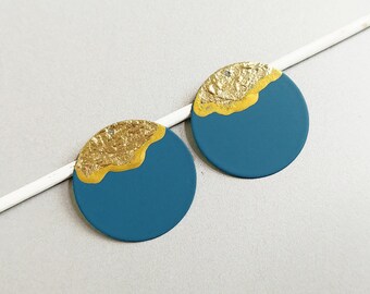 May include: Two teal blue, round earring components with a gold leaf texture on the top half. The gold leaf is separated from the blue by a thin gold line. The earrings are shown on a white stick.