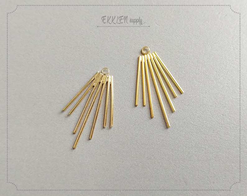 May include: Two gold-colored metal earring components. Each component features a small loop at the top and several straight, radiating bars. The components are arranged on a light gray surface. The text "EKKLEN supply" is visible.