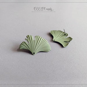 May include: Two sage green Ginkgo leaf-shaped stud earrings. The earrings have a textured, fan-like design with a post for pierced ears. The earrings are displayed on a light gray surface. The text "EKKLEN supply" is in the upper right corner.