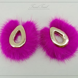 May include: A pair of pink faux fur earrings with gold metal teardrop-shaped accents. The earrings are approximately 5 cm in diameter.