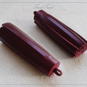 Peut inclure: Deux pendentifs en forme de glands de couleur bordeaux avec une extrémité en boucle pour la suspension. Les glands sont faits de fines bandes de tissu pliées et cousues ensemble.