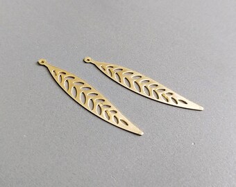 May include: Two gold-colored, leaf-shaped pendants with intricate cut-out designs. Each pendant has a small loop at the top for attaching to a chain or earring hook. The pendants are displayed on a gray surface.