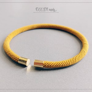 Può includere: Braccialetto in corda giallo senape con terminali color oro. Il braccialetto ha una forma circolare e presenta un design a corda ritorta e strutturata. Il testo "EKKLEN supply" è nell'angolo in alto a destra.