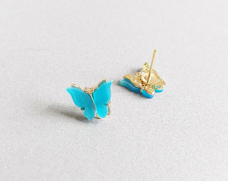 May include: A pair of butterfly stud earrings. The butterfly wings are a vibrant turquoise color, set in a gold-colored metal frame. The earrings have a post backing for pierced ears.