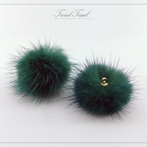 May include: Two dark green fluffy pom-poms with a gold metal loop on one. The pom-poms are on a white background.