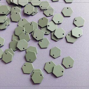 May include: A collection of small, hexagonal, light gray metal charms. Each charm has a single hole near the top for stringing or attaching. The charms are scattered across a light purple surface.