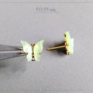 May include: Two butterfly-shaped stud earrings. The earrings have a pale green, iridescent finish and gold-colored metal accents. One earring is held by tweezers, while the other is displayed on a gray surface. The earrings are delicate and detailed.