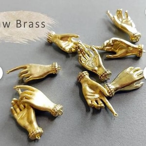 Op de afbeelding: Een verzameling kleine, goudkleurige handbedels. De bedels zijn gedetailleerd met vingers en polsen en hebben een lus om aan sieraden of andere knutselwerken te bevestigen. De tekst "Raw Brass" staat in de linkerbovenhoek.