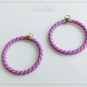 Op de afbeelding: Twee roze en blauwe gevlochten touwringen met gouden metalen sluitingen. De ringen hebben een diameter van ongeveer 7,5 cm.