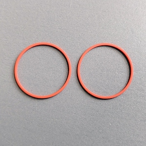 Puede incluir: Dos juntas tóricas rojas de goma sobre un fondo gris. Los anillos circulares son idénticos en tamaño y forma, con un acabado liso y mate. La imagen es un primer plano, que destaca los detalles del material de goma.