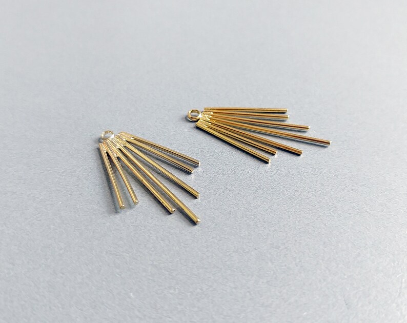 May include: Two gold-colored metal earring components. Each component features a small loop at the top and a fan-like arrangement of thin, straight bars radiating outwards. The components are displayed against a light gray background.