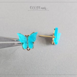 May include: A pair of butterfly stud earrings. The butterfly wings are a vibrant turquoise color with gold-colored trim. The earring posts are gold-colored. The earrings are displayed on a light gray surface.