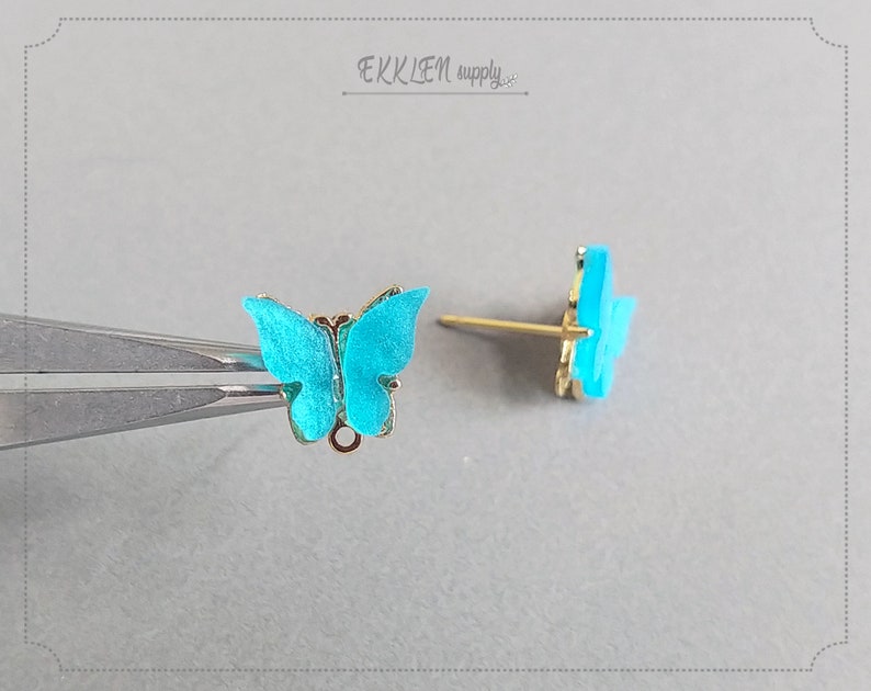 May include: A pair of butterfly stud earrings. The butterfly wings are a vibrant turquoise color with gold-colored trim. The earring posts are gold-colored. The earrings are displayed on a light gray surface.