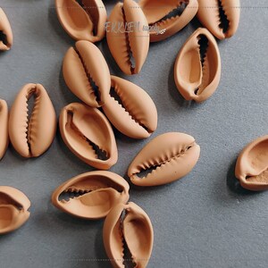 Puede incluir: Una colección de cuentas de concha de cauri de color marrón claro. Las conchas tienen un exterior liso y redondeado y una abertura dentada. Estas conchas decorativas son ideales para la fabricación de joyas y proyectos de artesanía.