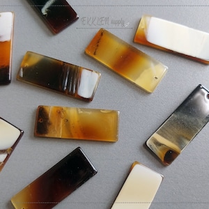 Peut inclure: Plusieurs pendentifs rectangulaires aux tons de marron, ambre et blanc, avec une finition brillante. Chaque pendentif est percé d'un petit trou à une extrémité. Les pendentifs sont disposés sur une surface grise. Le texte "EKKLEN supply" est visible.