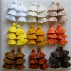 May include: Twelve pairs of multi-colored tassel earrings. Each pair has three layers of tassels. The colors are white, beige, yellow, orange, brown, and dark brown.