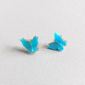May include: Two turquoise butterfly stud earrings with gold-colored accents. The earrings are small and delicate, with a bright, eye-catching color. The butterfly design is detailed and elegant, perfect for adding a touch of whimsy to any outfit.