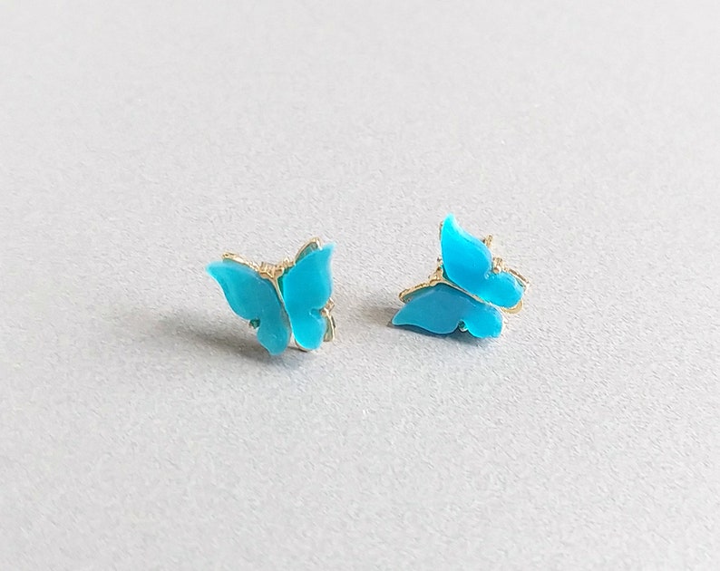 May include: Two turquoise butterfly stud earrings with gold-colored accents. The earrings are small and delicate, with a bright, eye-catching color. The butterfly design is detailed and elegant, perfect for adding a touch of whimsy to any outfit.