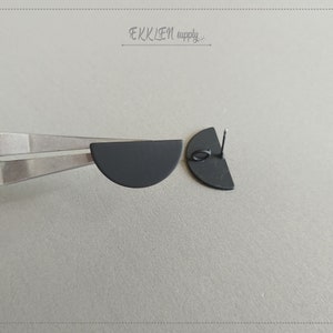 Pode incluir: Um par de brincos de semicírculo preto fosco. Um brinco é segurado por um alicate prateado, o outro está à direita. Os brincos têm um pino e um pequeno laço para fixação. O texto "EKKLEN supply" está no canto superior direito.