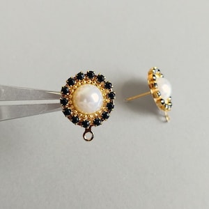 Puede incluir: Dos pendientes de botón dorados. Cada pendiente presenta una perla blanca central rodeada por un anillo de pequeñas piedras negras. Los pendientes tienen un poste y un pequeño lazo en la parte inferior. Sobre un fondo gris claro.