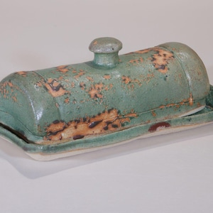 May include: A handmade ceramic butter dish, comprising a lid and a base. The dish is a muted teal with brown and orange speckles. The lid features a small, rounded knob. The base is rectangular with gently curved edges.